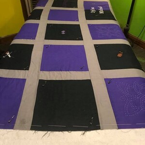 May include: A quilt top with a grid pattern of purple and black fabric squares. The squares are sewn together with gray fabric strips. The quilt top is pinned to a white surface. The quilt top has the names "Dylan" and "Macall" embroidered on it.