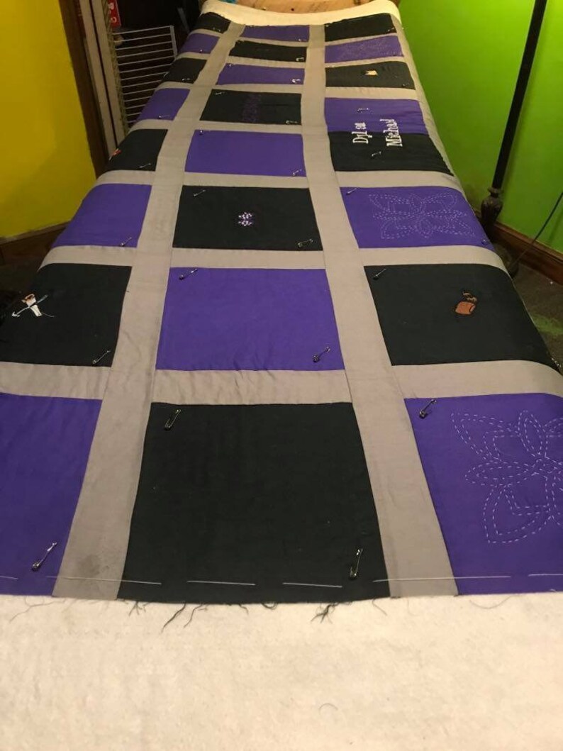 May include: A quilt top with a grid pattern of purple and black fabric squares. The squares are sewn together with gray fabric strips. The quilt top is pinned to a white surface. The quilt top has the names "Dylan" and "Macall" embroidered on it.