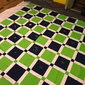 May include: A green and navy blue quilt with white squares. The quilt is pinned together and has various embroidered designs on each square. Some of the designs include a baseball, a basketball, a soccer ball, a football, a cross, and a heart.