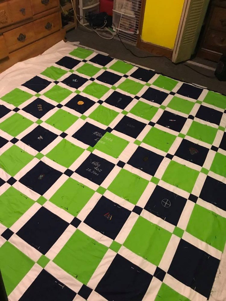 May include: A green and navy blue quilt with white squares. The quilt is pinned together and has various embroidered designs on each square. Some of the designs include a baseball, a basketball, a soccer ball, a football, a cross, and a heart.