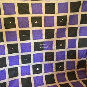 May include: A purple and black quilt with a white border. The quilt is made up of squares with different designs embroidered on them. The quilt has the text "Dylan Michael 3lbs 13in" embroidered on it.