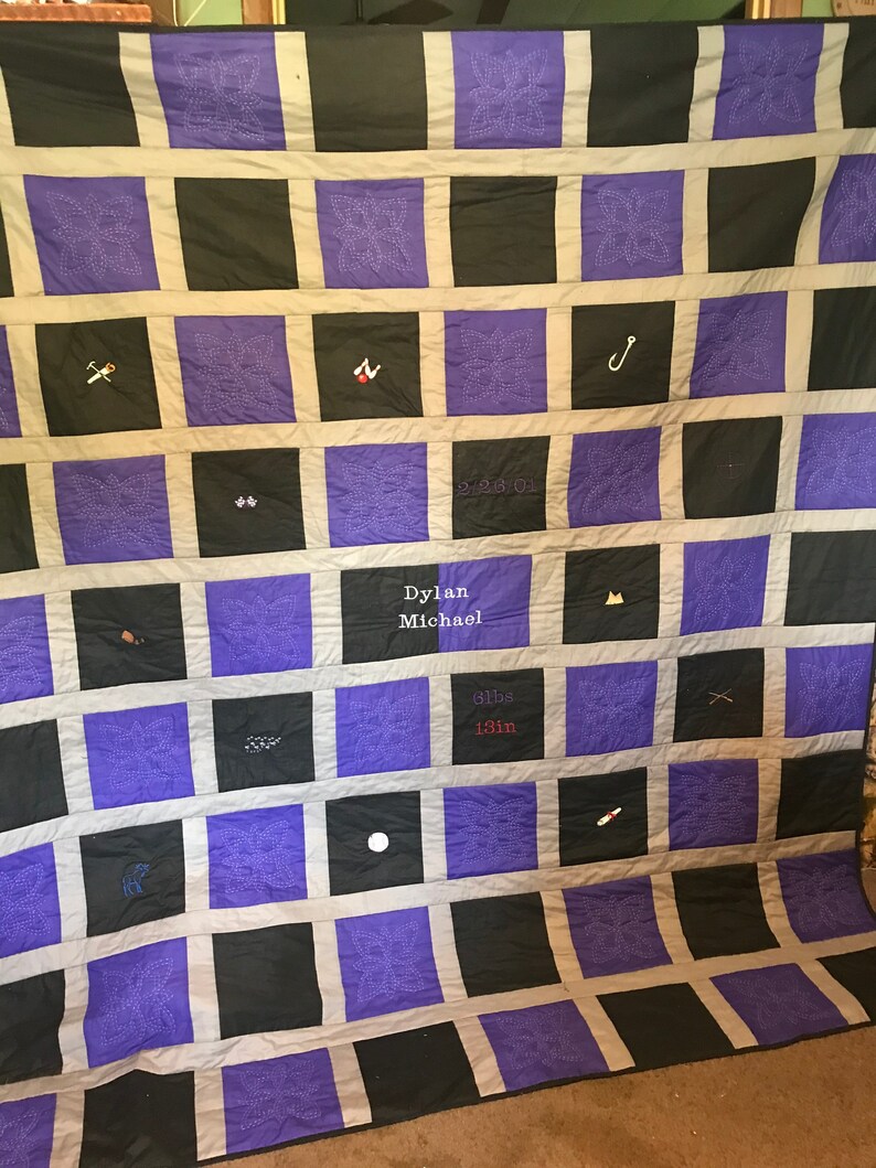 May include: A purple and black quilt with a white border. The quilt is made up of squares with different designs embroidered on them. The quilt has the text "Dylan Michael 3lbs 13in" embroidered on it.