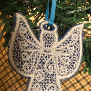 May include: White lace angel ornament with blue trim, hanging from a blue ribbon on a green artificial Christmas tree.