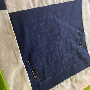 May include: A close-up of a quilt block with a navy blue fabric square in the center. The square has a white chalk outline of a maze pattern. The fabric is pinned to a white fabric background with gold pins.