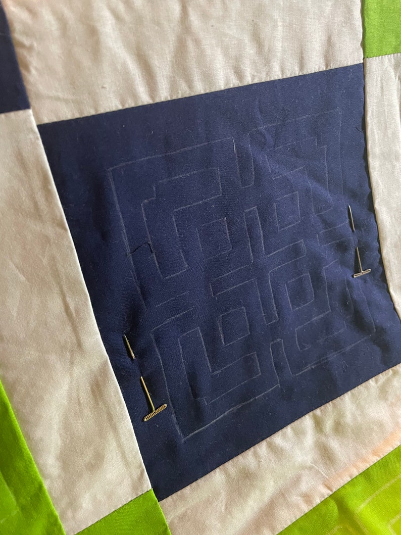 May include: A close-up of a quilt block with a navy blue fabric square in the center. The square has a white chalk outline of a maze pattern. The fabric is pinned to a white fabric background with gold pins.