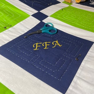 May include: A blue and white quilt with a green border. The quilt has a gold embroidered letter "FFA" in the center. There are sewing pins and a pair of scissors on the quilt.