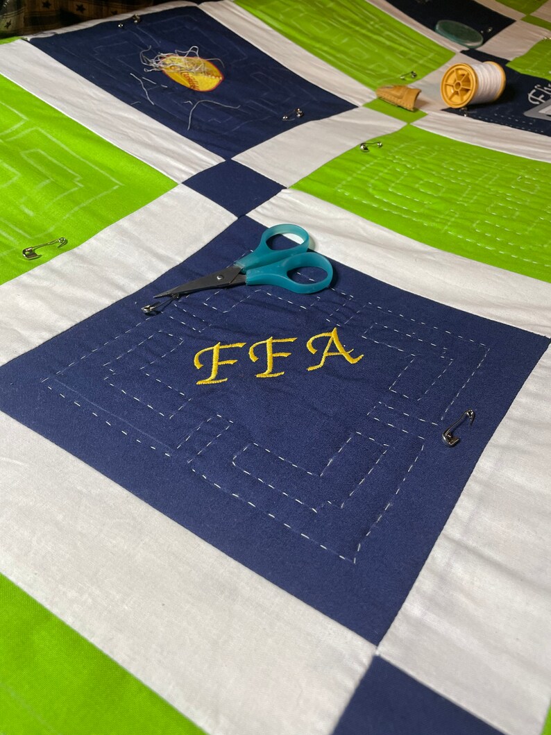 May include: A blue and white quilt with a green border. The quilt has a gold embroidered letter "FFA" in the center. There are sewing pins and a pair of scissors on the quilt.