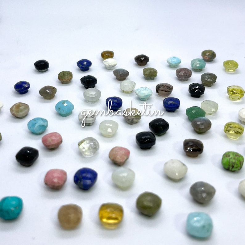 May include: Assortment of colorful gemstone cabochons in various shapes and sizes. The gemstones are displayed on a white surface.