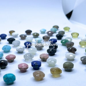 May include: Assortment of small, faceted gemstone cabochons in various colors, including turquoise, blue, green, pink, white, and black. The gemstones are arranged on a white surface. The text "gembasketin" is visible in the center of the image.