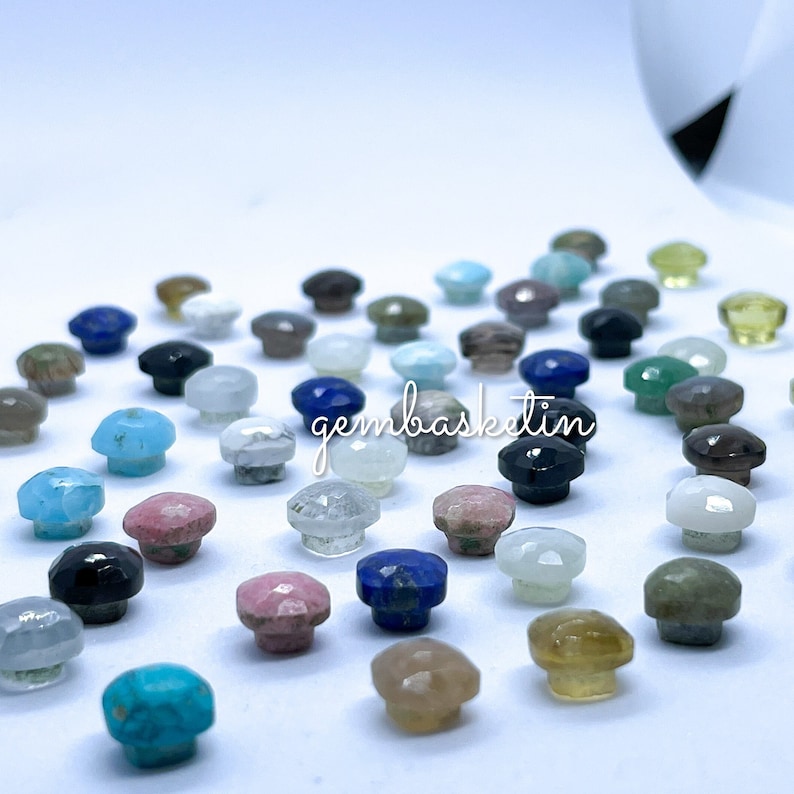 May include: Assortment of small, polished gemstone cabochons in various colors and shapes. The cabochons are arranged on a white surface. The text "gembasketlin" is visible in the image.