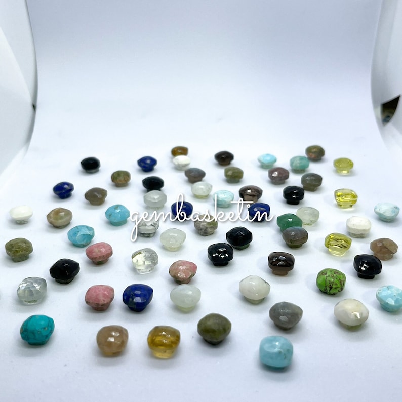 May include: Assortment of colorful gemstone cabochons in various shapes and sizes. The cabochons are displayed on a white surface.