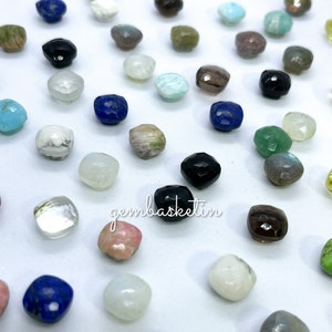May include: Assortment of faceted gemstone beads in various colors and shapes, including blue, green, black, white, and brown. The beads are arranged on a white surface. The text "gembaskeetin" is visible in the image.
