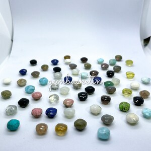 May include: Assortment of small, faceted gemstone cabochons in various colors, including turquoise, green, blue, pink, brown, black, and white. The gemstones are arranged on a white surface. The text "gembasketin" is visible in the center of the image.