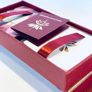 May include: A burgundy box with a gold lotus design and the text "Thoughtful Reflections" on a smaller burgundy card inside. The box is open and the lid is propped up, revealing a white card with a gold lotus design and a burgundy ribbon.