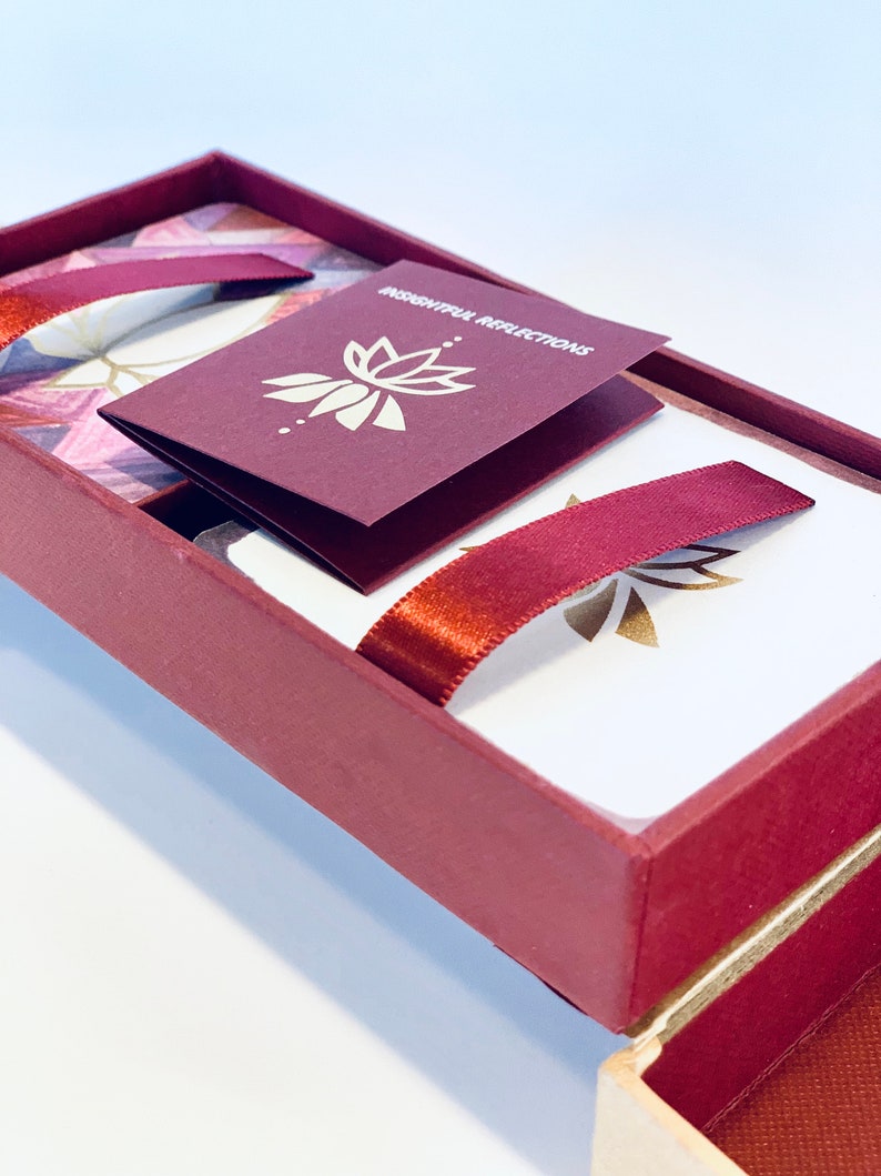 May include: A burgundy box with a gold lotus design and the text "Thoughtful Reflections" on a smaller burgundy card inside. The box is open and the lid is propped up, revealing a white card with a gold lotus design and a burgundy ribbon.