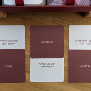 May include: Three white cards with maroon colored backs. The top of each card has a question in black text. The bottom of each card has a word in black text. The first card reads "Where in my life can I grow?" and "Family". The second card reads "Confidence" and "What step can I take today?" The third card reads "What have I gained from this?" and "Creativity".