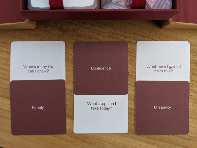 May include: Three white cards with maroon colored backs. The top of each card has a question in black text. The bottom of each card has a word in black text. The first card reads "Where in my life can I grow?" and "Family". The second card reads "Confidence" and "What step can I take today?" The third card reads "What have I gained from this?" and "Creativity".