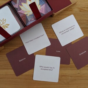 May include: A set of cards with white backgrounds and burgundy borders. The cards feature questions and words related to self-reflection and mindfulness. Some of the words on the cards include "Balance", "Love", "Gratitude", and "How have I created this?"