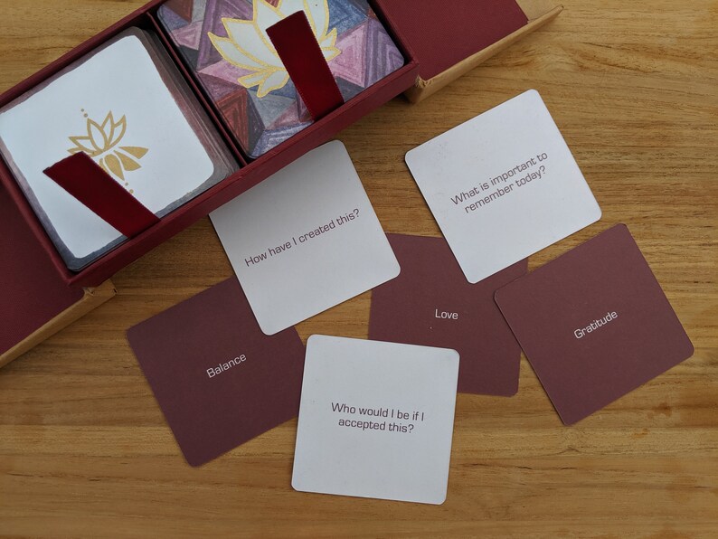 May include: A set of cards with white backgrounds and burgundy borders. The cards feature questions and words related to self-reflection and mindfulness. Some of the words on the cards include "Balance", "Love", "Gratitude", and "How have I created this?"