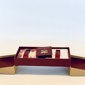 May include: A burgundy box with a gold and white lotus design on the lid. The box is open and the lid is propped up, revealing a set of cards with a white background and a burgundy ribbon.