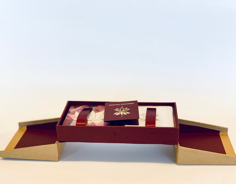 May include: A burgundy box with a gold and white lotus design on the lid. The box is open and the lid is propped up, revealing a set of cards with a white background and a burgundy ribbon.
