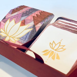 May include: A set of cards with a geometric pattern and a white lotus flower design in gold. The cards are in a burgundy box with a gold trim.