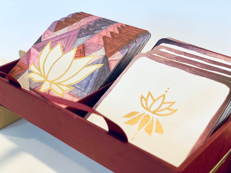 May include: A set of cards with a geometric pattern and a white lotus flower design in gold. The cards are in a burgundy box with a gold trim.