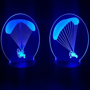 May include: Two blue LED night lights shaped like a paraglider with a person in the harness. The lights are on and illuminating the background.