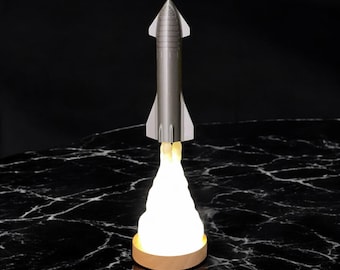 May include: A silver rocket ship shaped night light with a white light emanating from the bottom, resembling a flame. The light is on a wooden base and is sitting on a black marble surface.