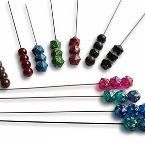 Könnte beinhalten: Eine Auswahl an farbenfrohen Würfeln und Perlensets auf dünnen Metallstäben. Die Würfel sind in verschiedenen Farben erhältlich, darunter Blau, Grün, Rot, Schwarz und Transparent. Die Perlen sind braun und rot. Die Würfel sind für Tischspiele.