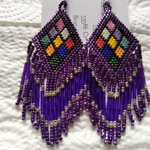 May include: Purple beaded earrings with a colorful geometric pattern and fringe. The earrings are handmade and have silver hooks.