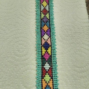 May include: A turquoise beaded bracelet with a colorful geometric pattern. The bracelet is made with small, round beads in various colors, including red, yellow, green, blue, and purple. The pattern is created by arranging the beads in a repeating design of diamonds and squares.