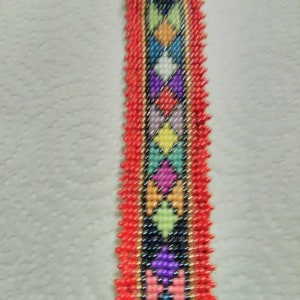 May include: A beaded bracelet with a central black band featuring a diamond pattern in various colors, including yellow, orange, blue, and purple. The edges are trimmed with red beads, and the overall design is intricate and colorful.