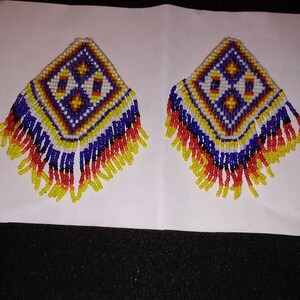May include: Two beaded earrings with a geometric design. The earrings are square-shaped with a diamond pattern in the center, featuring white, yellow, blue, and red beads. The bottom edges have beaded fringe in yellow, red, and blue.