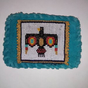 May include: A beaded Native American design of a black bird with red, yellow, and blue accents on a white background. The design is framed with gold beads and set on a turquoise blue fabric.