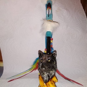 May include: A black wolf head pipe with a yellow and brown base. The pipe has a long stem with blue and white beads and feathers.