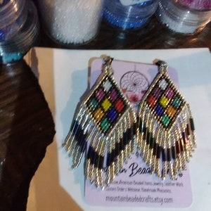 May include: A pair of colorful beaded earrings with a black and silver fringe. The earrings are in the shape of a diamond and have a variety of colors, including red, yellow, blue, green, and purple. The earrings are handmade and are available for purchase on the website mountainbeadedcrafts.etsy.com.