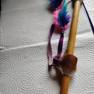 May include: A wooden flute with a fur-covered end and colorful feathers. The feathers are pink, teal, and purple, attached with a purple ribbon. The flute is made of light brown wood.