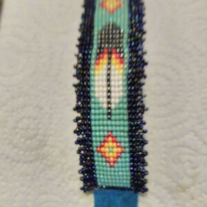 May include: A beaded bracelet with a turquoise background and a colorful feather design. The bracelet has a dark blue fringe on the edges.
