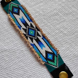 May include: A beaded cuff bracelet with a geometric design in shades of blue, green, black, and white. The edges are trimmed with gold beads, and the bracelet has a green suede base with black velvet accents and a gold snap closure.