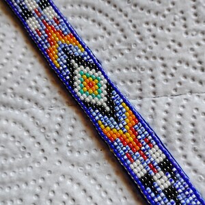 K&ouml;nnte beinhalten: Ein Armband aus Perlen mit blauem Hintergrund und bunten geometrischen Mustern. Das Armband hat ein zentrales Design mit einem wei&szlig;en, blauen, gelben und orangefarbenen Kreis.