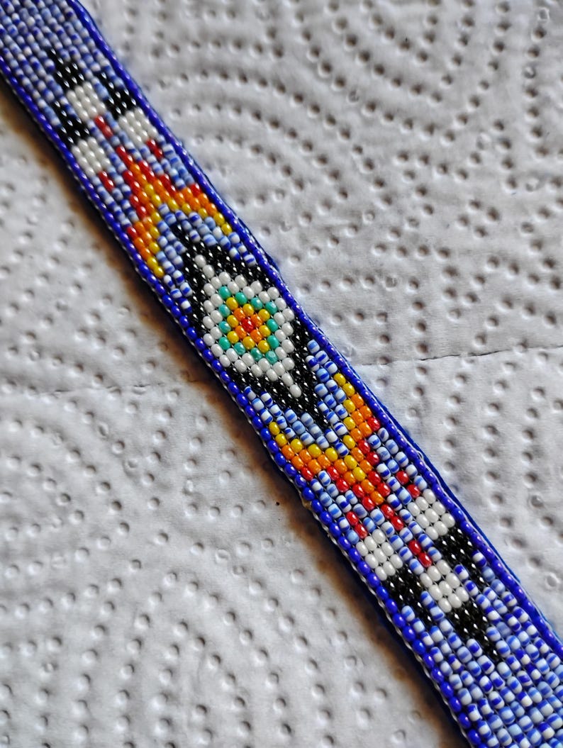 K&ouml;nnte beinhalten: Ein Armband aus Perlen mit blauem Hintergrund und bunten geometrischen Mustern. Das Armband hat ein zentrales Design mit einem wei&szlig;en, blauen, gelben und orangefarbenen Kreis.