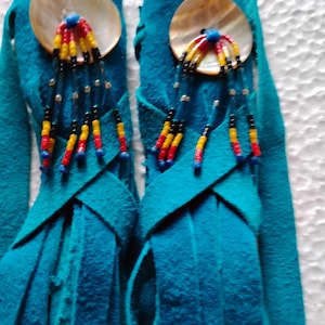 May include: Turquoise suede earrings with a fringe design. Each earring features a large, round, iridescent shell accent with beaded details in red, yellow, and black. The earrings have a long fringe.