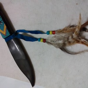 May include: A beaded knife with a blue, yellow, and red handle. The knife has a brown leather strap and feathers attached to it.