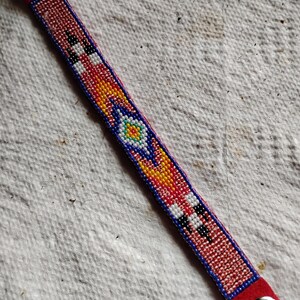 K&ouml;nnte beinhalten: Ein rotes Perlenarmband mit einem geometrischen Muster in Rot-, Wei&szlig;-, Blau-, Gelb- und Gr&uuml;nt&ouml;nen. Das Armband hat Lederenden mit Metallschnallen.