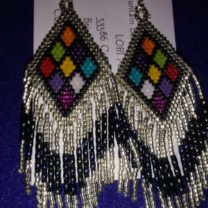 May include: A pair of beaded earrings with a black and silver fringe. The earrings feature a diamond-shaped design with colorful beads in shades of purple, turquoise, green, yellow, orange, and red.