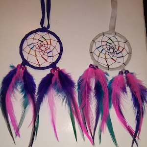 May include: Two dream catchers with colorful feathers. One has a dark purple hoop and ribbon, the other a white hoop and ribbon. Both have rainbow-colored beadwork in the center and pink, purple, and teal feathers.