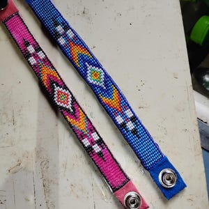 K&ouml;nnte beinhalten: Zwei Perlenarmb&auml;nder mit bunten geometrischen Mustern. Das Armband links ist pink mit wei&szlig;en und gelben Akzenten. Das Armband rechts ist blau mit wei&szlig;en und gelben Akzenten. Beide Armb&auml;nder haben einen Druckknopfverschluss.