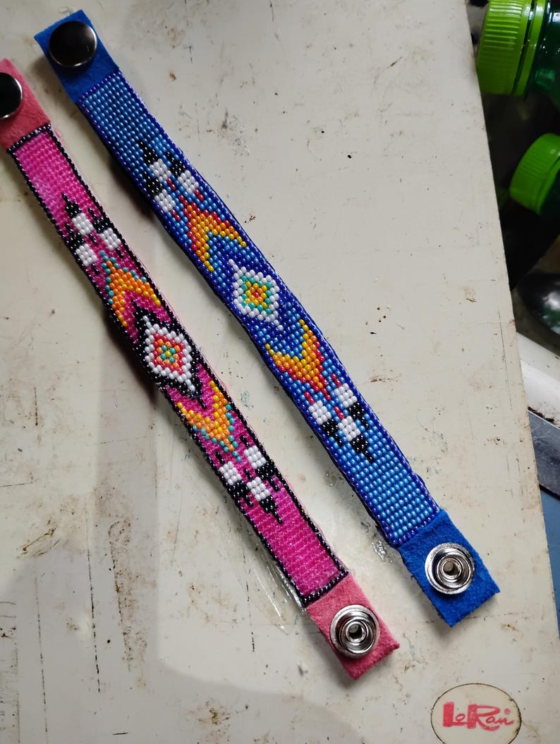 K&ouml;nnte beinhalten: Zwei Perlenarmb&auml;nder mit bunten geometrischen Mustern. Das Armband links ist pink mit wei&szlig;en und gelben Akzenten. Das Armband rechts ist blau mit wei&szlig;en und gelben Akzenten. Beide Armb&auml;nder haben einen Druckknopfverschluss.