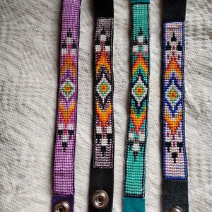 K&ouml;nnte beinhalten: Vier Perlenarmb&auml;nder mit bunten geometrischen Mustern und Lederriemen mit Druckkn&ouml;pfen. Die Armb&auml;nder sind in den Farben Lila, T&uuml;rkis, Schwarz und Wei&szlig;.